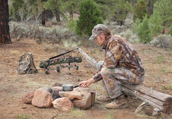 modern bowhunter sitting alone on log in camp and cooking over campfire in the woods