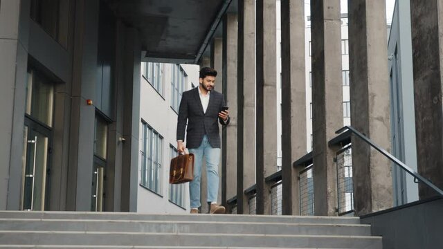 Full length portrait of arabian man walking down on stairs with modern smartphone in one hand and leather brown suitcase in another. Concept of business, success and confidence. Modern gadget for work