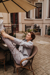Positive woman in suit stretches while sitting on street cafe. Active young lady in grey jacket and pants smiles and has fun outside