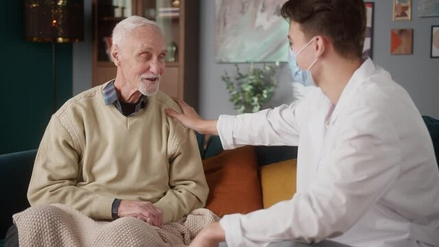 Young doctor in mask consulting elderly old man, telling about medical appointment, sitting on sofa. Retirement and pension, pensioner speaking with male nurse, supporting during coronavirus. 
