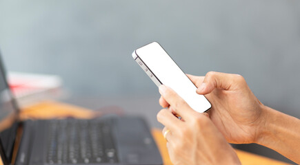 Hand holding black mobile phone with white screen mockup while working with labtop at home office