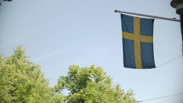 Swedish flag hanging from building with trees in the background in a sunny day in Sweden. Filmed handheld in 25fps