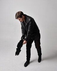 Full length portrait of a  brunette man wearing leather jacket  and holding a science fiction gun.  Standing  action pose isolated  against a grey studio background.