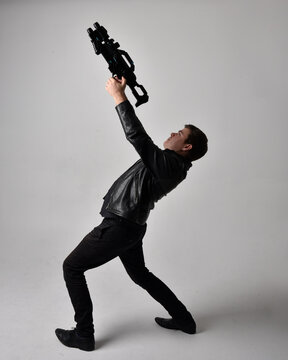 Full length portrait of a  brunette man wearing leather jacket  and holding a science fiction gun.  Standing  action pose isolated  against a grey studio background.