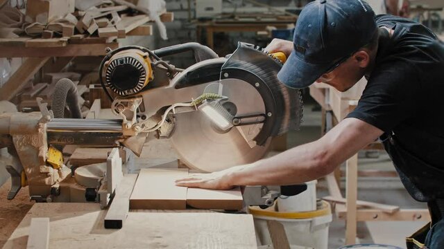 Man in a carpentry workshop cutting a small pieces of the wooden detail using a big circular saw