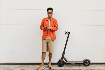 Happy young curly dark-skinned man in orange jacket, beige shorts and colorful t-shirt smiles,...