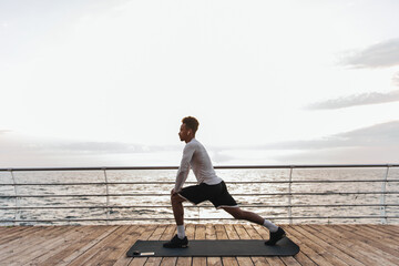 Curly dark-skinned man in black shorts and white sport t-shirt listens to music in headphones and stretches on yoga mat near sea.