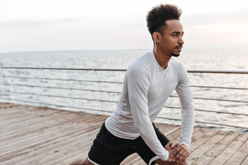 Handsome curly brunette man in black shorts and long-sleeved t-shirt stretches outside. Dark-skinned guy works out near sea.