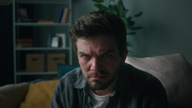 Angry man portrait, brutal male person with beard frowning, sitting on sofa in living room, looking in camera with furious view. Feeling irritation and anger.