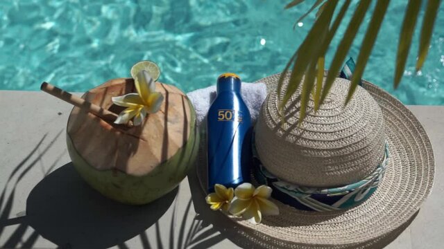 Video footage of green young coconut close up with bamboo straw, bottle of sunscreen SPF 50, sun hat, tropical yellow flower frangipani, palm shade on the edge of swimming pool in Bali