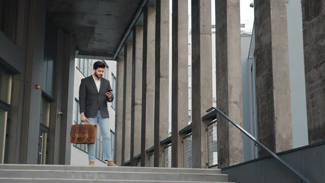 Handsome arabian man dressed in stylish formal clothes standing on stairs with suitcase and smartphone in hands. Modern gadget for work. Business concept. Arabian businessman standing on stairs
