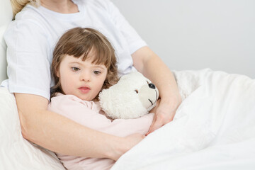 Mom hugging a girl with down syndrome holding a teddy bear at home in the bedroom on the bed. Ordinary childcare in a family for children with disabilities