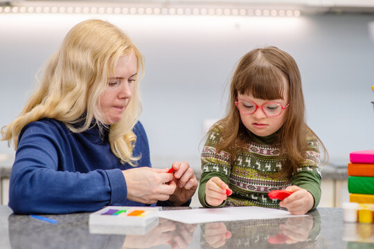 A girl with down syndrome sits next to her mother and is engaged in creativity, drawing a picture in an album. Education for people with disabilities concept