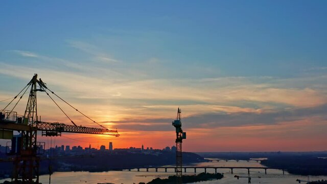 Aerial vertical view of delightful sunset against background of high tower cranes on construction site and beautiful cityscape with bridges across wide city river