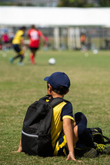 Junior soccer player rests with his teammates on a sideline waiting for their next match in a football school tournament, while some pitches still in action.