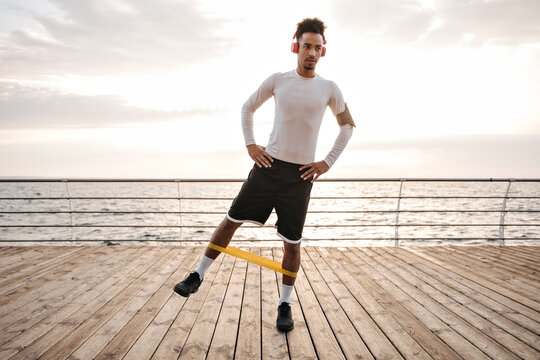 Dark-skinned curly brunette man in white long-sleeved t-shirt and black shorts listens to music in headphones and does exercises with fitness rubber near sea.