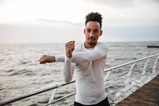 Charming dark-skinned man in white sport long-sleeved shirt stretches near sea. Young guy working out at beach.