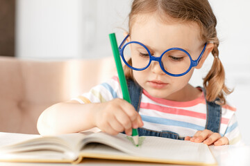 Smart child girl with glasses draws colored pencils at table in white kitchen. Developing children's skills, kid hobbies, education and learning, back to school. Background, copy space