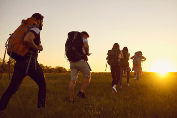 Group of tourists hikers hiking with backpacks in green summer field at sunset during summer...