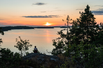 Beautiful sunset on the Baltic Sea. Young woman sitting on top of high stone cliff and watches the setting sun. Bright golden orange pink tones of the sunset sky. Islands of the Stockholm archipelago.