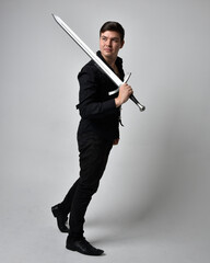 Full length portrait of a  brunette man wearing black shirt and gothic waistcoat holding sword.  Standing  action pose isolated  against a grey studio background.
