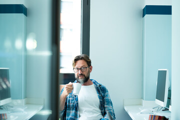 Fototapeta premium Mature man portrait looking on camera while drink coffee at office - freelance online job with computer concept lifestyle - adult caucasian people in coworking space - blue mood colors modern people