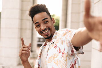 Excited bearded charming man in white floral summer shirt points up, looks into camera, smiles and...