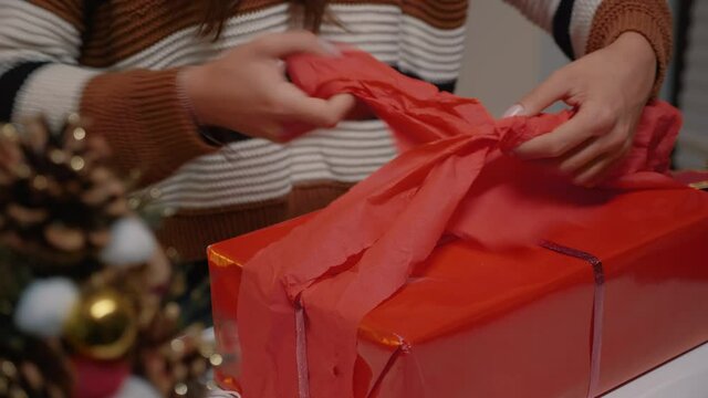 Young adult wrapping gift box in red paper to give to friends at christmas eve celebration party. Woman decorating present with ribbon for winter season joyful tradition in december