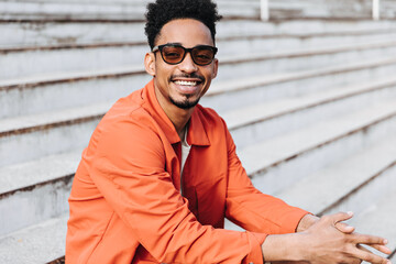 Cheerful good-humored brunet man in sunglasses and orange jacket smiles sincerely and sits on...