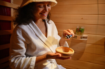 Smiling woman pouring few drops of basil essential oil into wooden mortar while relaxing in wooden sauna