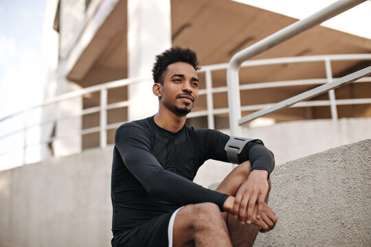 Portrait of young curly bearded dark-skinned man in black long-sleeved t-shirt and shorts sitting on stairs outside.