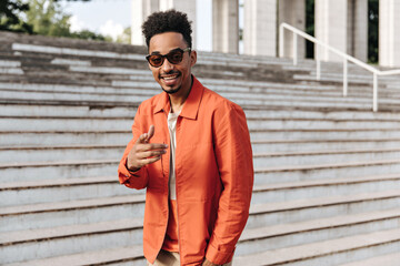 Cool cheerful brunet dark-skinned man j stylish sunglasses and orange jacket points at camera and poses near stairs outdoors.