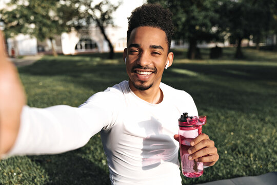 Attractive young curly dark-skinned man in white sport t-shirt smiles sincerely, holds pink water bottle and takes selfie in park.