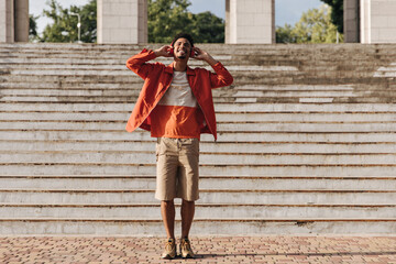 Excited optimistic brunet man in beige shorts and orange jacket smiles sincerely, listens to music...