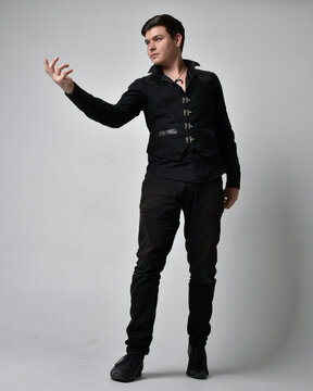 Full length portrait of a brunette man wearing black shirt and gothic waistcoat.  Standing pose isolated  against a grey studio background.