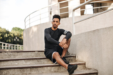 Brown-eyed brunet dark-skinned bearded man in long-sleeved t-shirt and shorts sits on stairs outside and looks away.