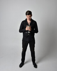 Full length portrait of a brunette man wearing black shirt and gothic waistcoat.  Standing pose isolated  against a grey studio background.