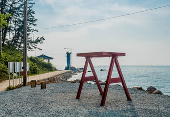 Wooden frame for park swing on gravel lot overlooking ocean water with lighthouse in background..