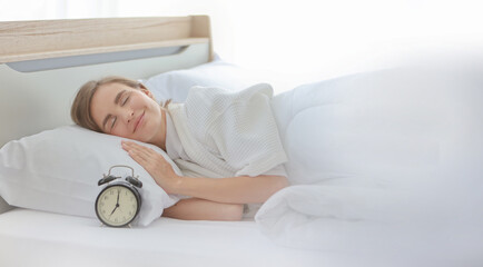 Cheerful and healthy caucasian young woman wearing pajama try waking up with alarm clock on the bed with sunlight