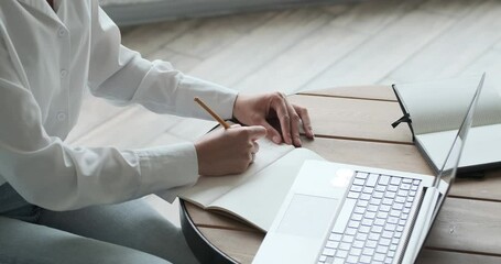 Close up view of young woman writing in notepad and looking at laptop with tutorial. Student doing homework preparing for exams and learn foreign language using video call with teacher.