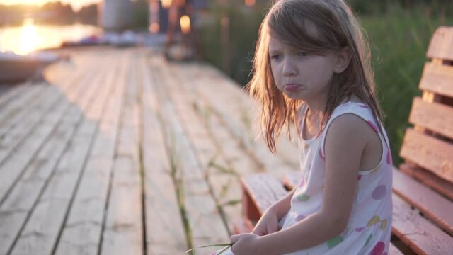 A sad little girl sits by the river at sunset in the summer.
