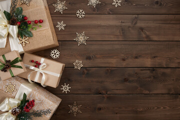 Rustic Christmas background with border of gift boxes wrapped in kraft paper decorated with red berries and Christmas greenery on dark wood desk. Xmas, winter, holiday concept. Flat lay, copy space
