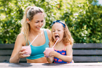 A beautiful girl and her little daughter drink a refreshing smoothie while sitting on a bench in the garden