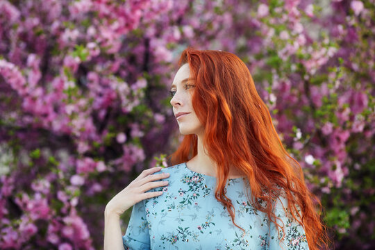 Stylish russian beauty with long red loose hair smiling and looking into the distance. Sensual portrait of a girl posing against a background of blurred pink flowers