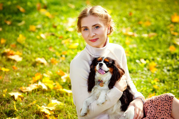 A young blonde woman in a white sweater is sitting on the grass of an autumn sunny park and hugs her Cavalier King Charles Spaniel dog. A woman and her pet in nature.