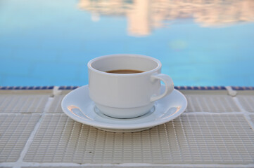 A cup of coffee on a saucer stands in the shade on the background of the pool