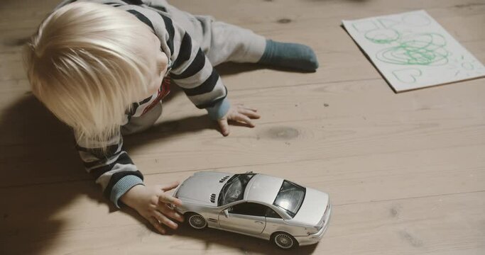 View of blonde baby boy playing silver car model on the wooden floor. Room filled with morning sunlight. Tiny fingers with toys.