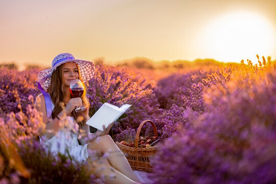 young beautiful attractive lady in white dress with white hat reads a book and holds a glass of red wine in lavender field during sunrise