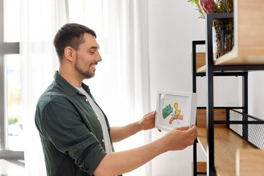 home improvement, decoration and people concept - happy smiling man placing watercolor picture in frame to shelf