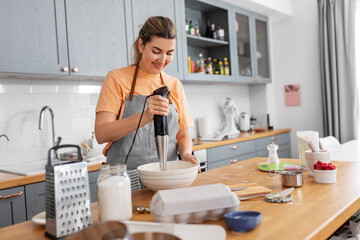 culinary, baking and people concept - happy smiling young woman cooking food on kitchen at home and using immersion blender
